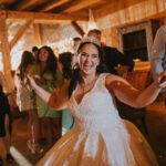 Eine fröhliche Braut in einem weißen Kleid und einer Tiara tanzt nach ihrer standesamtlichen Trauung mit den Gästen bei einem rustikalen Empfang mit Holzbalken. Sie lächelt breit und hat die Arme ausgestreckt. Menschen in festlicher Kleidung umgeben sie und warmes, stimmungsvolles Licht verstärkt die festliche Atmosphäre. - Elis Wedding - Hochzeitsfotograf Frankfurt