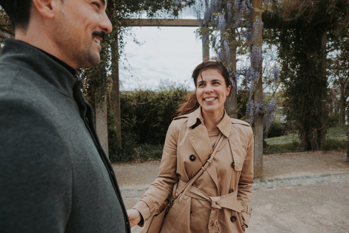Eine Frau in einem hellbraunen Trenchcoat lächelt und sieht einen Mann in einer dunklen Jacke an. Sie stehen draußen auf einem Feldweg mit Holzbögen, die mit Kletterpflanzen und violetten Blumen im Hintergrund geschmückt sind, was die ungezwungene Freundlichkeit zweier Menschen suggeriert, die sich vielleicht mitten in Kennenlernen befinden. - Elis Wedding - Hochzeitsfotograf Frankfurt
