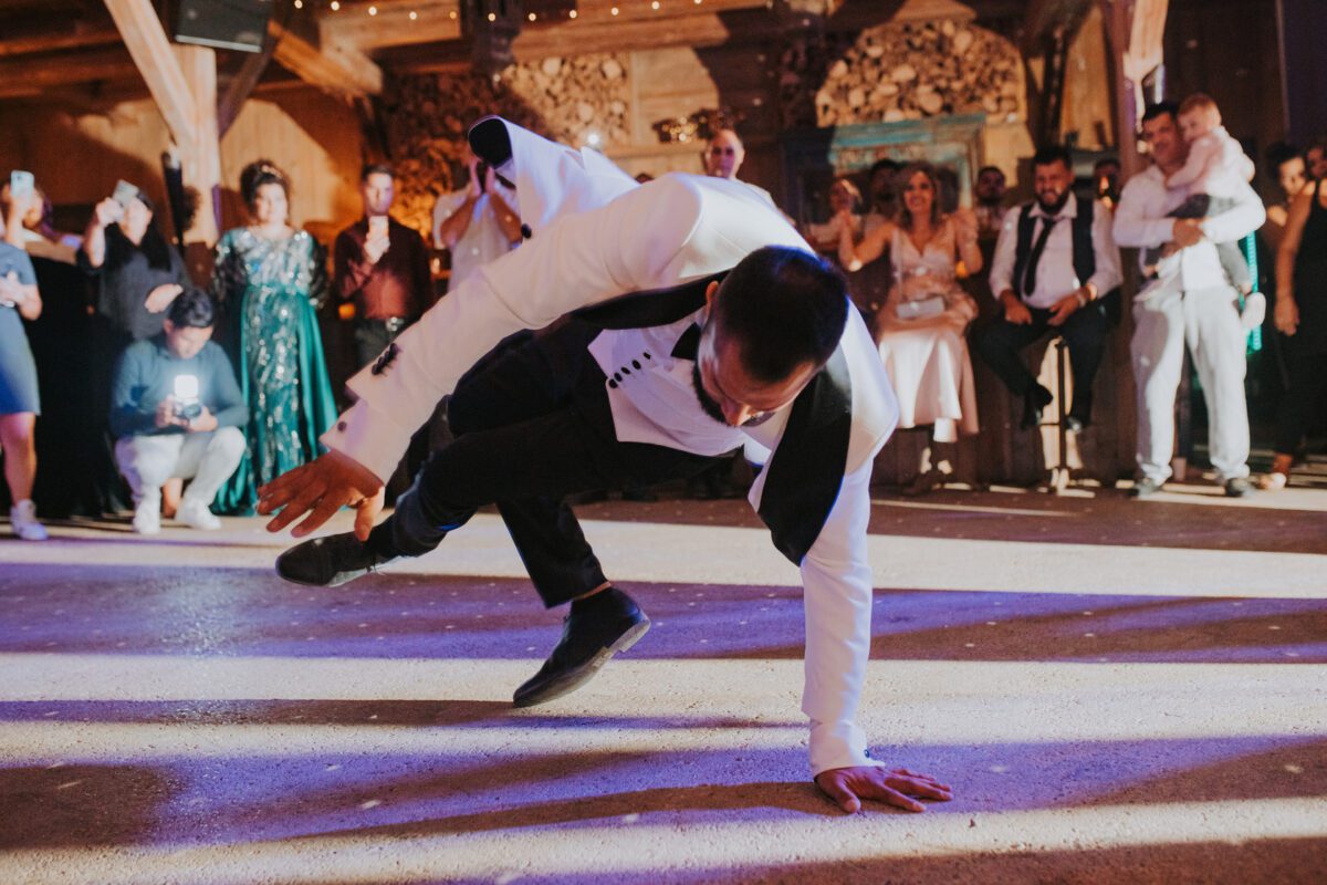 Ein Mann in formeller Kleidung, darunter eine weiße Jacke und schwarze Hosen, führt einen dynamischen Breakdance-Move aus und balanciert dabei auf einer Hand in der Mitte eines Partysaals. Gäste in eleganter Kleidung schauen im Hintergrund zu und jubeln, einige halten den Moment mit ihren Handys fest. Ein Hochzeitsfotograf ist damit beschäftigt, die lebhafte Szene in warmer Beleuchtung zu fotografieren. - Elis Wedding - Hochzeitsfotograf Frankfurt