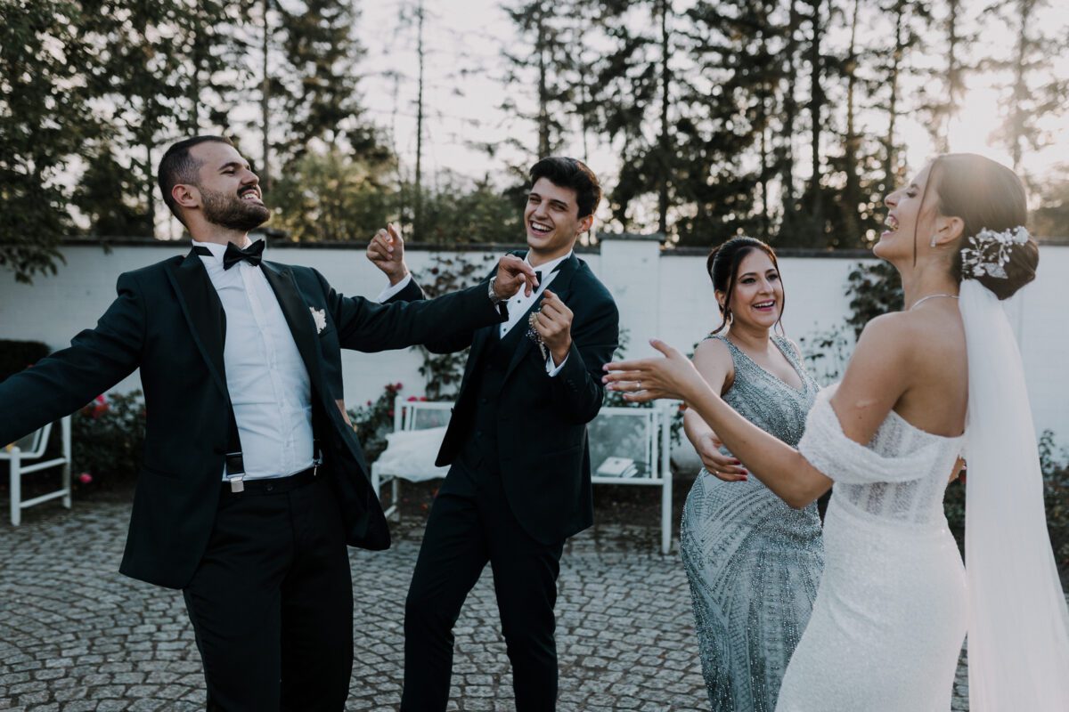 Vier formell gekleidete Personen vergnügen sich draußen, tanzen und lachen. Der Bräutigam in schwarzem Smoking und Fliege und die Braut in einem weißen Kleid mit schulterfreien Trägern und floralem Haarschmuck genießen ihre standesamtliche Trauung. Zwei elegant gekleidete Gäste gesellen sich vor einer Kulisse aus Bäumen und Sitzgelegenheiten im Freien zu ihnen. - Elis Wedding - Hochzeitsfotograf Frankfurt