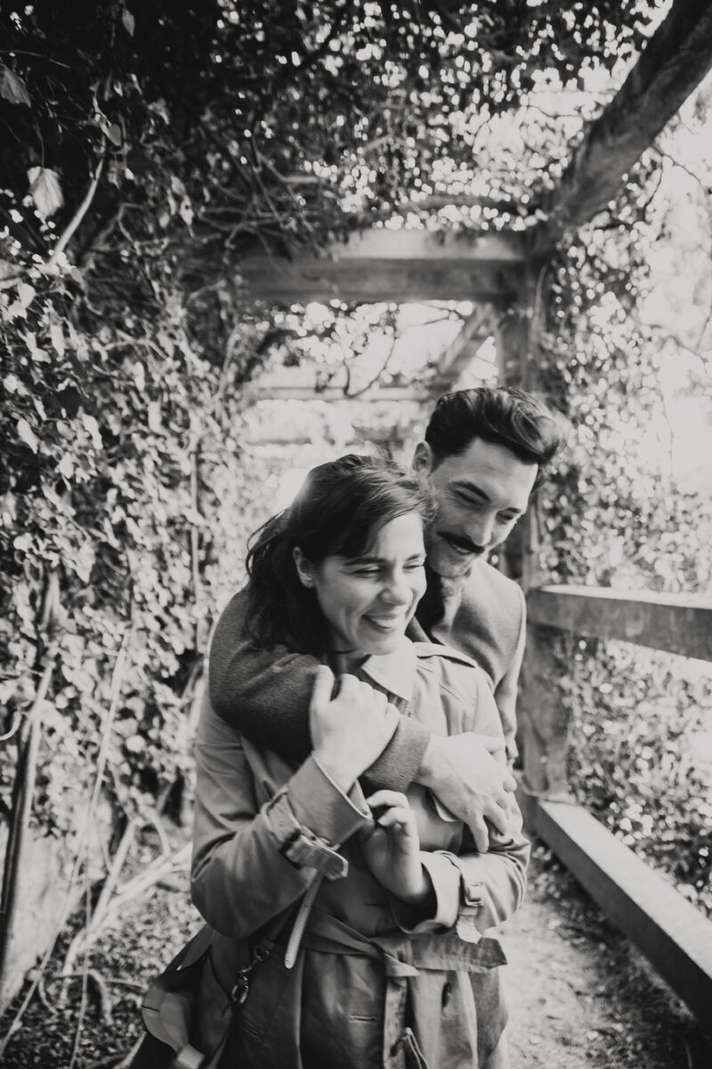 Ein Schwarzweißfoto aus einem Fotoshooting zeigt ein lächelndes Paar, das im Freien unter einer mit Weinreben bewachsenen Pergola spazieren geht. Der Mann steht hinter der Frau und legt liebevoll seine Arme um ihre Schultern. Beide wirken zufrieden und glücklich und genießen einen friedlichen Moment zusammen. - Elis Wedding - Hochzeitsfotograf Frankfurt