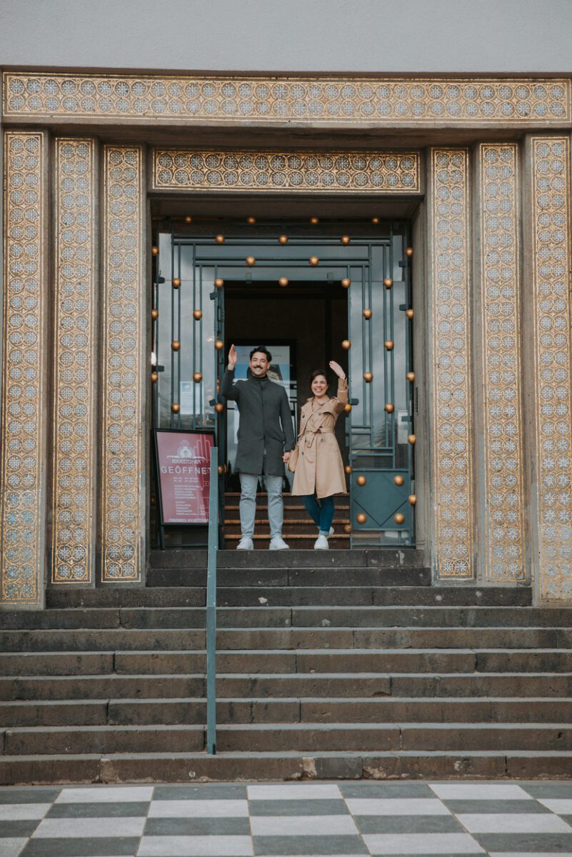 Ein Mann und eine Frau stehen am Eingang eines Gebäudes mit kunstvollen goldenen und blauen Mosaikdetails. Der Mann trägt einen grauen Mantel und eine graue Hose, während die Frau einen hellbraunen Trenchcoat trägt. Beide lächeln und winken und stehen auf einer grauen Treppe mit einem schwarz-weiß karierten Boden davor, perfekt für ein spontanes Fotoshooting. - Elis Wedding - Hochzeitsfotograf Frankfurt