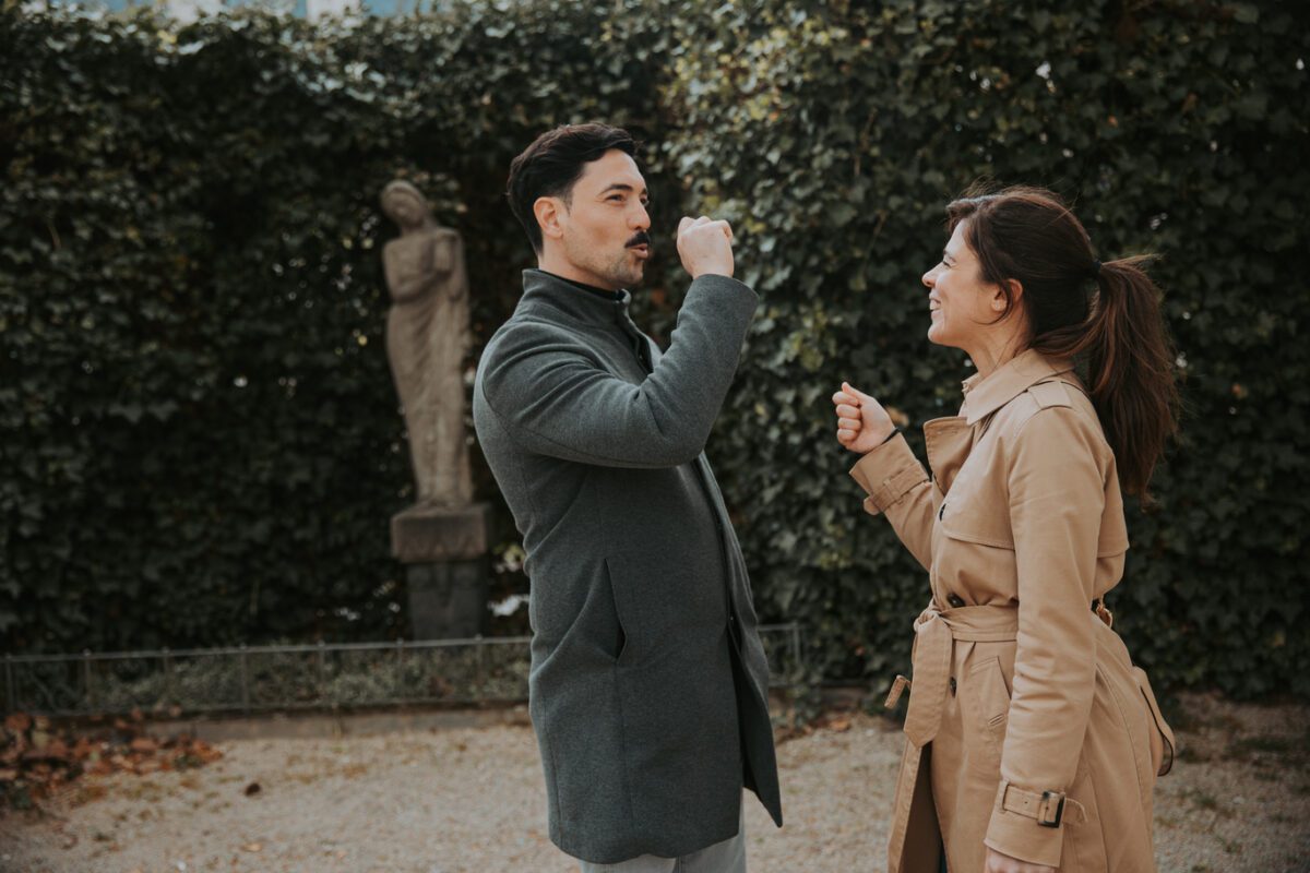 Ein Mann und eine Frau stehen draußen vor einer Hecke mit Blättern und einer Steinstatue. Der Mann im grauen Mantel und die Frau im beigen Trenchcoat scheinen während einer Art Fotoshooting zu tanzen oder spielerisch aufeinander zu gestikulieren. Sie lächeln und genießen einen unbeschwerten Moment miteinander. - Elis Wedding - Hochzeitsfotograf Frankfurt