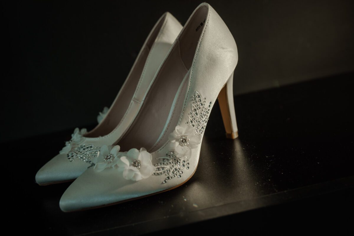 Ein Paar elegante weiße Stöckelschuhe auf dunklem Untergrund, perfekt für eine standesamtliche Trauung in Frankfurt. Die Schuhe sind mit dekorativen Blumen und aufwendigen Perlen an den Seiten verziert, was ihnen einen Hauch von Glanz verleiht. Der Hintergrund ist dunkel, wodurch die Schuhe als Blickfang hervorgehoben werden. Die Beleuchtung betont ihren Glanz und ihre Details. - Elis Wedding - Hochzeitsfotograf Frankfurt