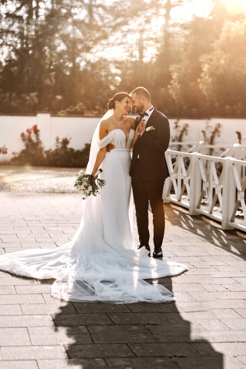 Braut und Bräutigam stehen eng beieinander und umarmen sich in einer sonnigen Umgebung im Freien. Die Braut trägt ein schulterfreies weißes Kleid mit langer Schleppe und hält einen Blumenstrauß in der Hand. Der Bräutigam, in schwarzem Anzug und Fliege, lächelt sie liebevoll an. Ein weißer Zaun und Grün rahmen diesen wunderschönen Moment ein, der von ihrem Hochzeitsfotografen festgehalten wurde. - Elis Wedding - Hochzeitsfotograf Frankfurt