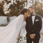 Braut und Bräutigam lächeln sich auf einer sonnigen Brücke im Freien an. Die Braut, in einem weißen Kleid mit langem Schleier, hält einen Strauß weißer Blumen. Der Bräutigam, in einem dunklen Anzug mit einer weißen Ansteckblume, steht mit gefalteten Händen da. Im Hintergrund sind Bäume und ein weißer Zaun zu sehen – eine perfekte Szene für ihren Hochzeitsfotografen. - Elis Wedding - Hochzeitsfotograf Frankfurt