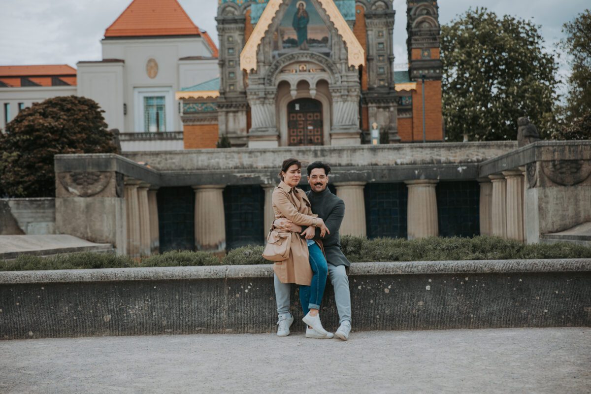 Ein Paar sitzt am Rand einer niedrigen Steinmauer. Die Frau trägt einen hellbraunen Trenchcoat, graue Hosen und weiße Schuhe, und der Mann trägt einen dunklen Mantel, blaue Jeans und weiße Turnschuhe. Hinter ihnen befindet sich ein kunstvolles Gebäude mit Säulen, Bögen und detaillierten Ornamenten, perfekt für ihr standesamtliches Trauungs-Fotoshooting in Frankfurt. - Elis Wedding - Hochzeitsfotograf Frankfurt