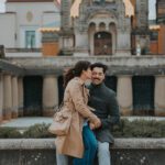 Eine Frau in einem hellbraunen Trenchcoat küsst einen lächelnden Mann in einem grauen Mantel auf die Wange. Sie sitzen auf einem Steinsims vor einem historischen Gebäude mit kunstvoller Architektur und Säulen. Die Frau trägt eine hellbraune Umhängetasche und sie sind von Grün umgeben, perfekt für ein spontanes Fotoshooting eines Hochzeitsfotografen. - Elis Wedding - Hochzeitsfotograf Frankfurt