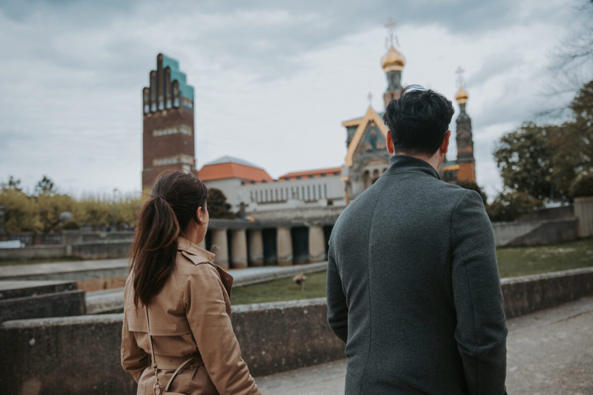 Ein Mann und eine Frau gehen Seite an Seite auf eine Kirche mit goldenen Kuppeln in einer parkähnlichen Umgebung in Frankfurt zu. Die Frau mit dem dunklen Haar, das zu einem Pferdeschwanz gebunden ist, trägt einen hellbraunen Trenchcoat, und der Mann mit dem kurzen dunklen Haar trägt einen dunkelgrauen Mantel. Unter einem bewölkten Himmel sind Bäume und moderne Gebäude zu sehen. - Elis Wedding - Hochzeitsfotograf Frankfurt