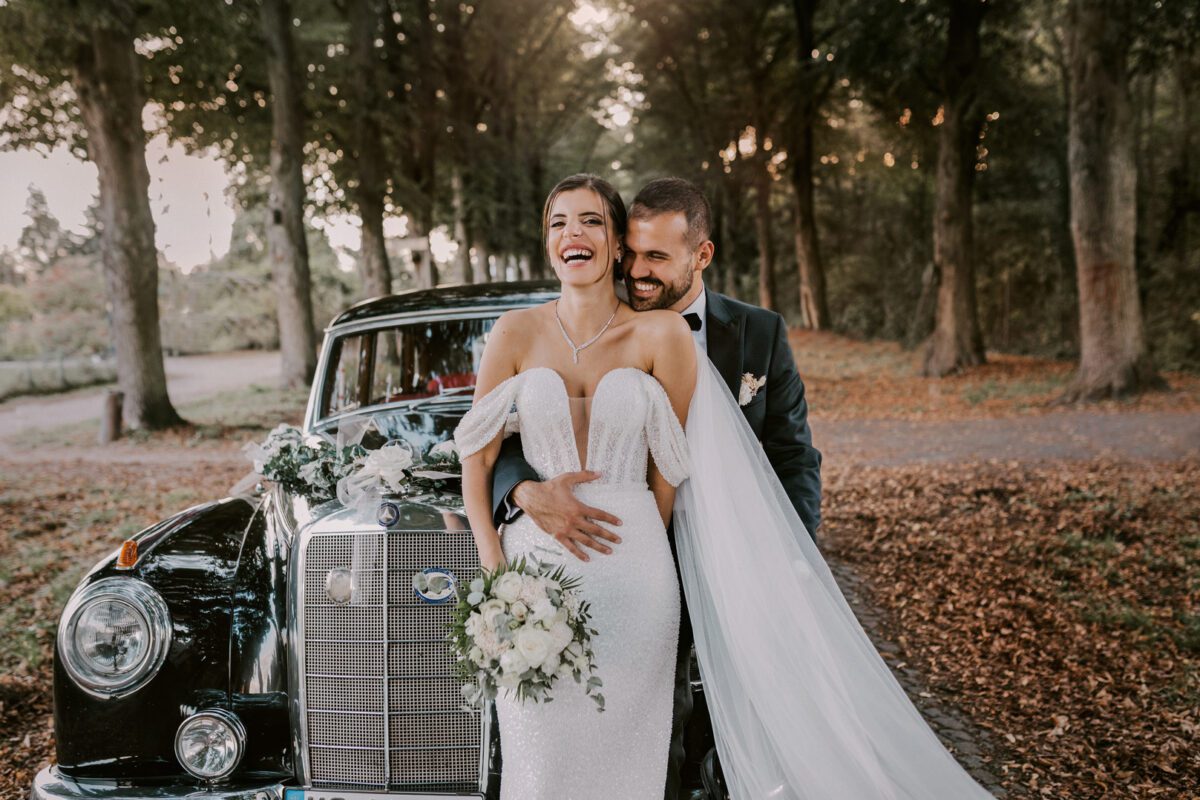 Eine Braut in einem weißen trägerlosen Brautkleid und einer Perlenkette lacht fröhlich, während der Bräutigam im schwarzen Smoking sie von hinten umarmt. Sie stehen vor einem mit Blumen geschmückten schwarzen Oldtimer auf einem von Bäumen gesäumten Weg, der mit gefallenen Blättern bedeckt ist. Die Szene, die ihr Hochzeitsfotograf eingefangen hat, ist voller Glück und Romantik. - Elis Wedding - Hochzeitsfotograf Frankfurt