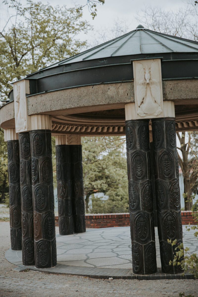 Ein runder, offener Pavillon verfügt über mehrere schwarze, kunstvoll geschnitzte Säulen mit kunstvollen Mustern. Die Struktur hat ein konisches, graues Dach und helle Reliefplatten an der Spitze jeder Säule. Er ist ideal für eine ruhige standesamtliche Trauung oder Hochzeitsfotografen-Sessions und ist von Laubbäumen und einem Steinweg umgeben, was der Szene eine natürliche Atmosphäre verleiht. - Elis Wedding - Hochzeitsfotograf Frankfurt