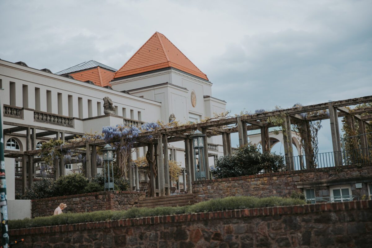 Ein großes, elegantes Gebäude mit weißen Wänden und einem roten Ziegeldach steht vor einem bewölkten Himmel in Frankfurt. Im Vordergrund erstreckt sich horizontal eine mit Weinreben und violetten Blumen geschmückte Holzpergola. Unterhalb der Pergola wertet eine mit Grünpflanzen gesäumte Backsteinmauer den Anblick auf, perfekt für eine standesamtliche Trauung oder ein Fotoshooting. - Elis Wedding - Hochzeitsfotograf Frankfurt