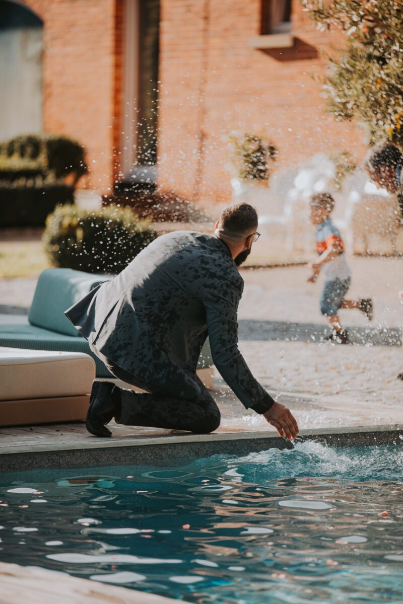 Ein Mann im nassen, grauen Anzug kniet am Rand eines Swimmingpools und spritzt mit seiner rechten Hand Wasser. Er trägt eine Brille und schwarze Schuhe. Im Hintergrund rennt ein Kind davon und schaut den Mann an. Die Kulisse ist ein Außenbereich mit Backsteingebäuden und Sträuchern – perfekt für Hochzeitsfotografie (Hochzeitsfotograf). - Elis Wedding - Hochzeitsfotograf Frankfurt