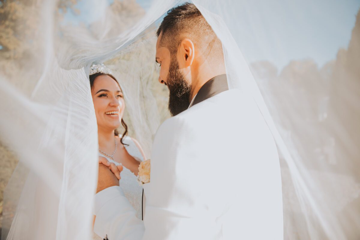 Ein Paar lächelt sich an seinem Hochzeitstag herzlich an. Die Braut, die ein weißes Kleid und eine Tiara trägt, hält die Hände des Bräutigams während ihrer standesamtlichen Trauung. Der Bräutigam, der einen Bart und einen weißen Anzug mit schwarzem Kragen trägt, blickt sie liebevoll an, während der Schleier sie teilweise umhüllt und eine sanfte, intime Atmosphäre schafft. - Elis Wedding - Hochzeitsfotograf Frankfurt