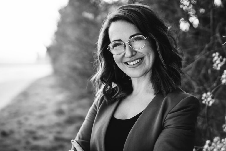 Ein Schwarz-Weiß-Porträt einer lächelnden Person mit welligem, schulterlangem Haar und Brille. Sie trägt einen Blazer über einem dunklen Oberteil und steht im Freien vor einem verschwommenen, von Bäumen gesäumten Hintergrund. Die Arme sind lässig verschränkt. Die Gesamtstimmung ist positiv und zuversichtlich. - Elis Wedding - Hochzeitsfotograf Frankfurt