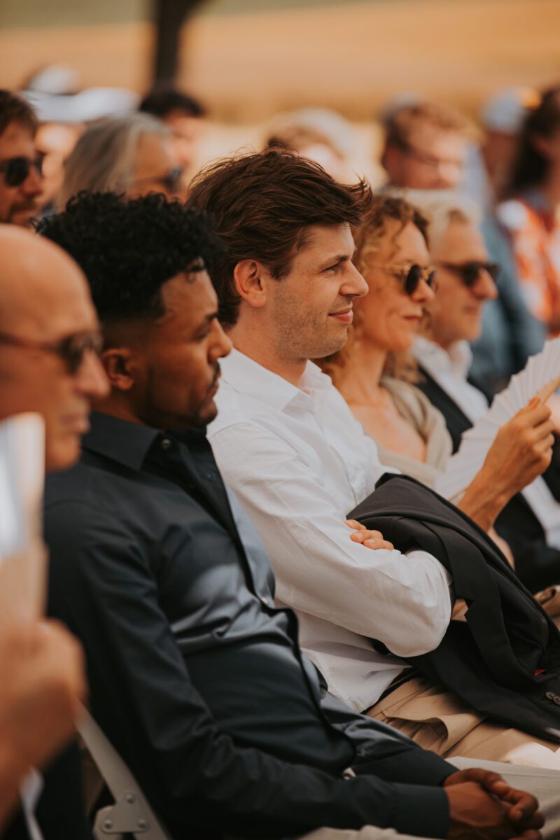 Eine Gruppe von Menschen sitzt im Freien und beobachtet aufmerksam eine Veranstaltung. Das Publikum ist leger gekleidet, einige halten Programme oder Papiere in den Händen. Das sonnige Ambiente verleiht der natürlichen, möglicherweise ländlichen Umgebung Wärme. Es scheint der ideale Moment für einen Frankfurter Hochzeitsfotografen zu sein, um die Freude und das Zusammensein aller einzufangen. - Elis Wedding - Hochzeitsfotograf Frankfurt