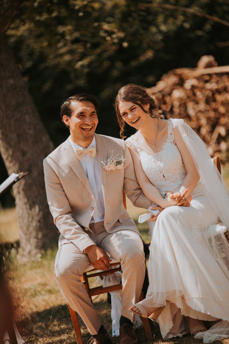 Ein Paar sitzt an seinem Hochzeitstag im Freien und teilt einen freudigen Moment, der von seinem Hochzeitsfotografen festgehalten wurde. Die Braut trägt ein weißes Spitzenkleid mit Schleier und der Bräutigam einen hellbeigen Anzug mit weißer Fliege. Sie halten sich an den Händen und lächeln breit. Im Hintergrund sitzen Bäume und gestapeltes Holz, was eine rustikale Umgebung suggeriert. - Elis Wedding - Hochzeitsfotograf Frankfurt
