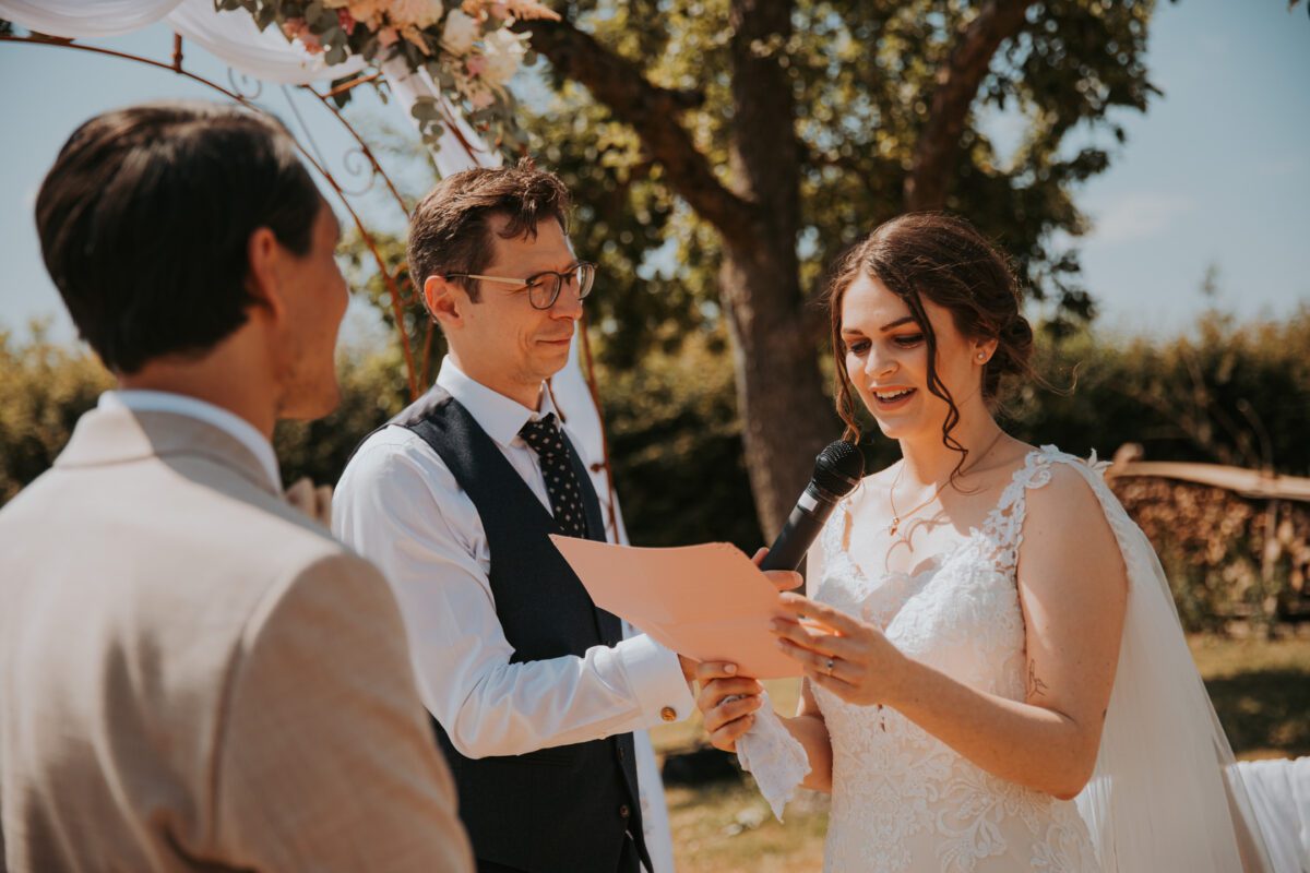 Die Braut im Spitzenbrautkleid liest ihre Gelübde von einem rosa Blatt Papier, hält ein Mikrofon und lächelt neben dem Bräutigam im beigen Anzug. Ein Geistlicher mit Brille und blauer Weste schaut zu und ist bereit zu assistieren. Sie stehen draußen unter einem mit Blumen geschmückten Torbogen, inmitten von Bäumen und Büschen in Frankfurt. - Elis Wedding - Hochzeitsfotograf Frankfurt