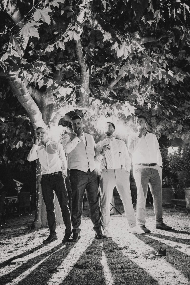 Eine Schwarz-Weiß-Fotografie zeigt vier Männer, die unter einem großen, mit Lichterketten geschmückten Baum stehen. Sie stehen in einer Reihe, jeder hält eine Zigarre in der Hand und ist halbformell gekleidet. Der mit Kies bedeckte Boden wirft Schatten vom Baum, die sich in Richtung Kamera erstrecken und ein Gefühl von Vertrautheit in Frankfurt hervorrufen. - Elis Wedding - Hochzeitsfotograf Frankfurt