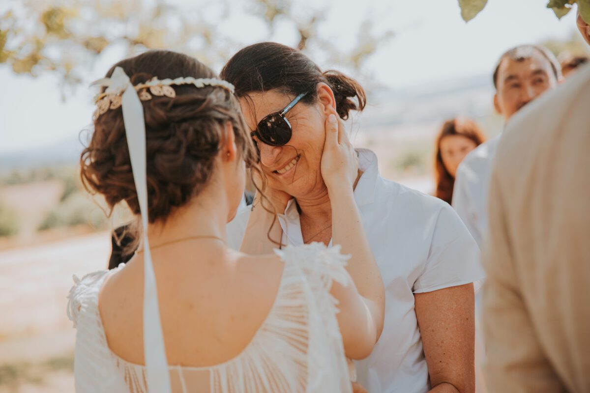 Eine Braut, von hinten zu sehen, trägt ein weißes Kleid und einen Blumenkopfschmuck, hält zärtlich das Gesicht einer Frau mit Sonnenbrille und weißer Bluse. Sie teilen einen innigen Moment unter einem Baum während ihrer standesamtlichen Trauung, mit verschwommenen Gästen im Hintergrund und einer malerischen Außenkulisse, die von ihrem Hochzeitsfotografen eingefangen wurde. - Elis Wedding - Hochzeitsfotograf Frankfurt
