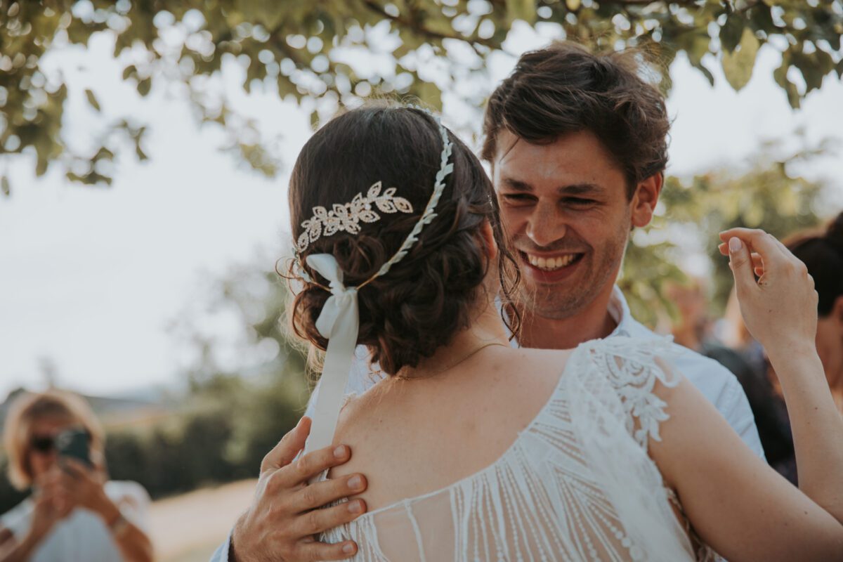 Braut und Bräutigam umarmen sich im Freien in Frankfurt. Der Bräutigam lächelt breit und hält die Braut, die von hinten zu sehen ist. Sie trägt ein zartes Spitzenkleid und ein Haarteil mit aufwendigen floralen Details. Im Hintergrund fängt ihr Hochzeitsfotograf den Moment inmitten einer natürlichen Kulisse ein. - Elis Wedding - Hochzeitsfotograf Frankfurt