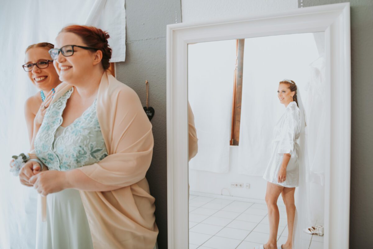 Zwei Frauen, eine in einem hellblauen Spitzenkleid und mit Brille, die andere mit Brille und beigem Schal, lächeln, während sie neben einem Ganzkörperspiegel stehen. In der Spiegelung sieht sich eine Frau in einem weißen Gewand lächelnd an. Im Hintergrund sind weiße Wände und hängende Stoffe zu sehen, die den freudigen Moment während ihrer standesamtlichen Trauung einfangen. - Elis Wedding - Hochzeitsfotograf Frankfurt