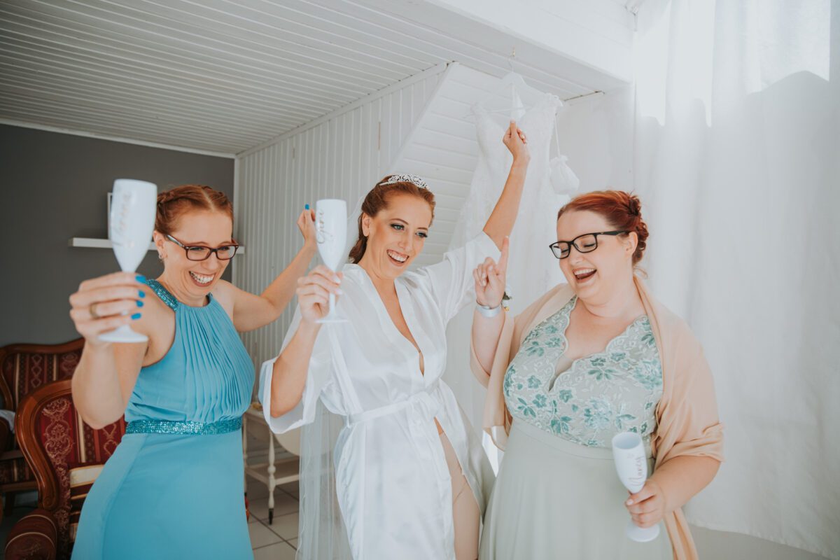 Drei Frauen feiern in einem hell erleuchteten Raum. Die Braut, die ein weißes Gewand und eine Tiara trägt, steht in der Mitte und hat die Arme erhoben, als würde sie für ein Fotoshooting posieren. Die beiden Brautjungfern, in blaue und grüne Kleider gekleidet, halten weiße Becher und freuen sich mit der Braut. Im Hintergrund hängt ein weißes Brautkleid. - Elis Wedding - Hochzeitsfotograf Frankfurt