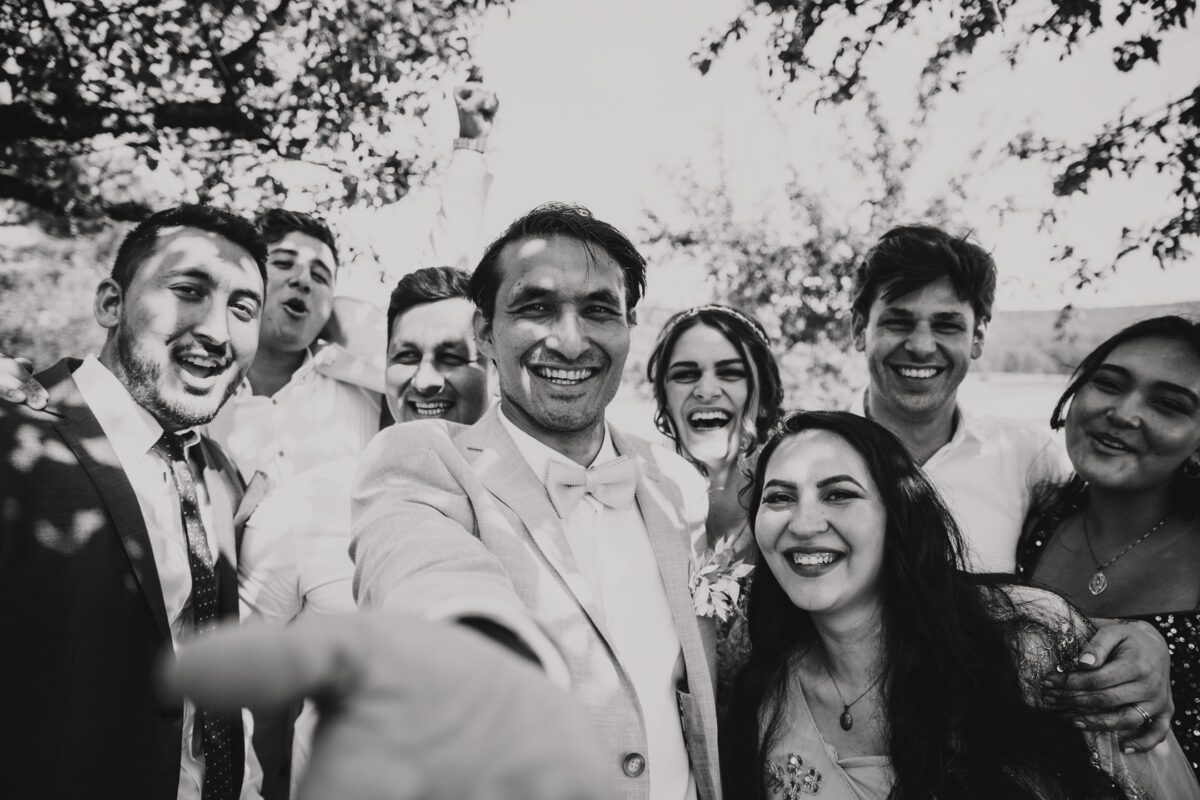 Ein Schwarzweißfoto zeigt eine fröhliche Gruppe von acht Menschen, die im Freien im gesprenkelten Licht der Blätter lächeln und lachen. Ein Mann in der Mitte, der einen hellen Anzug trägt, streckt seinen Arm in Richtung Kamera und schafft so eine dynamische und lebhafte Atmosphäre während eines Standesamtlichen Trauungs-Fotoshootings in Frankfurt. - Elis Wedding - Hochzeitsfotograf Frankfurt