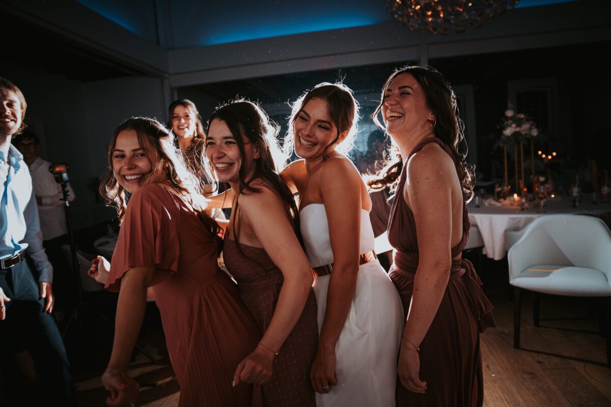 Vier fröhliche junge Frauen in Kleidern stehen dicht beieinander und lächeln in einem warm beleuchteten Raum, wahrscheinlich bei einer standesamtlichen Trauung oder einer Feier. Sie lachen und genießen den Moment, im Hintergrund sind ein runder Tisch und Stühle zu sehen. Der Raum verfügt über sanftes, stimmungsvolles Licht und einen Kronleuchter darüber, der an elegante Fotografie erinnert, die man oft in Frankfurt sieht. - Elis Wedding - Hochzeitsfotograf Frankfurt