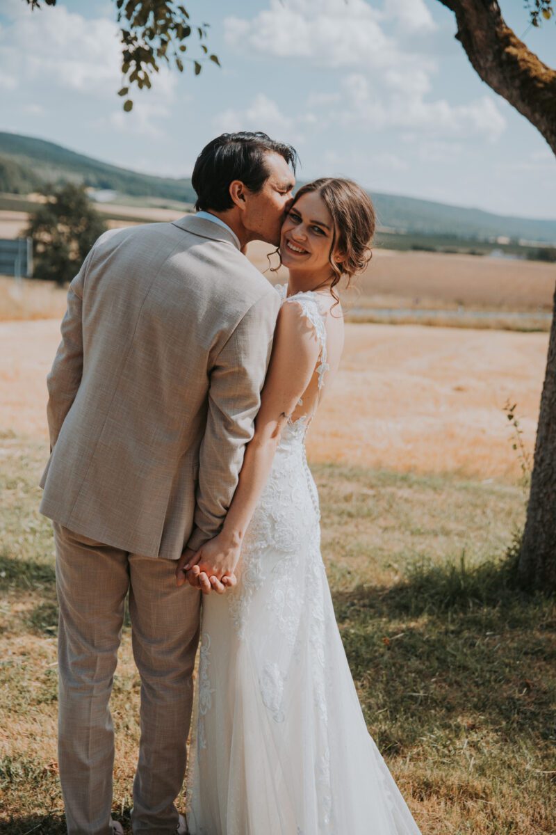Ein frisch verheiratetes Paar steht im Freien, hält Händchen und blickt in die Kamera, während eines gefühlvollen Fotoshootings. Der Bräutigam, in einem hellgrauen Anzug, beugt sich vor, um die lächelnde Braut auf die Schläfe zu küssen. Die Braut trägt ein weißes Spitzenkleid und blickt über ihre Schulter. Sie sind von Grün und einem offenen Feld mit einem blauen Himmel im Hintergrund eingerahmt. - Elis Wedding - Hochzeitsfotograf Frankfurt