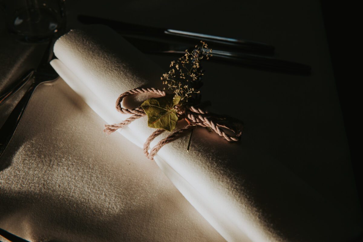 Eine weiße Stoffserviette ist gerollt und mit einem rosa Seil zusammengebunden, das mit einem kleinen Zweig getrockneter Blumen und einem grünen Blatt geschmückt ist. In der schwach beleuchteten Umgebung spielen Schatten auf der Serviette. Auf dem Tisch, der für das Fotoshooting bereitsteht, sind im Hintergrund Besteck, darunter ein Messer und eine Gabel, zu sehen. - Elis Wedding - Hochzeitsfotograf Frankfurt