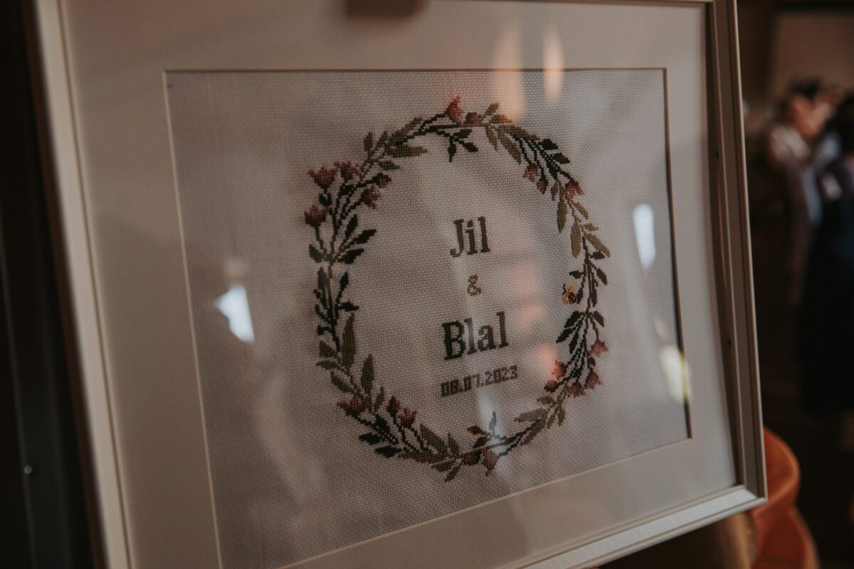 Eine gerahmte Stickerei zeigt in der Mitte die Namen „Jil & Bilal“, umgeben von einem runden Blumenkranz mit grünen Blättern und rosa Blüten. Unter ihren Namen ist in kleinerer Schrift das Datum „10.01.2023“ eingestickt. Dieses Stück ist perfekt für eine Hochzeit in Frankfurt und spiegelt die feine Kunstfertigkeit eines Hochzeitsfotografen wider. - Elis Wedding - Hochzeitsfotograf Frankfurt