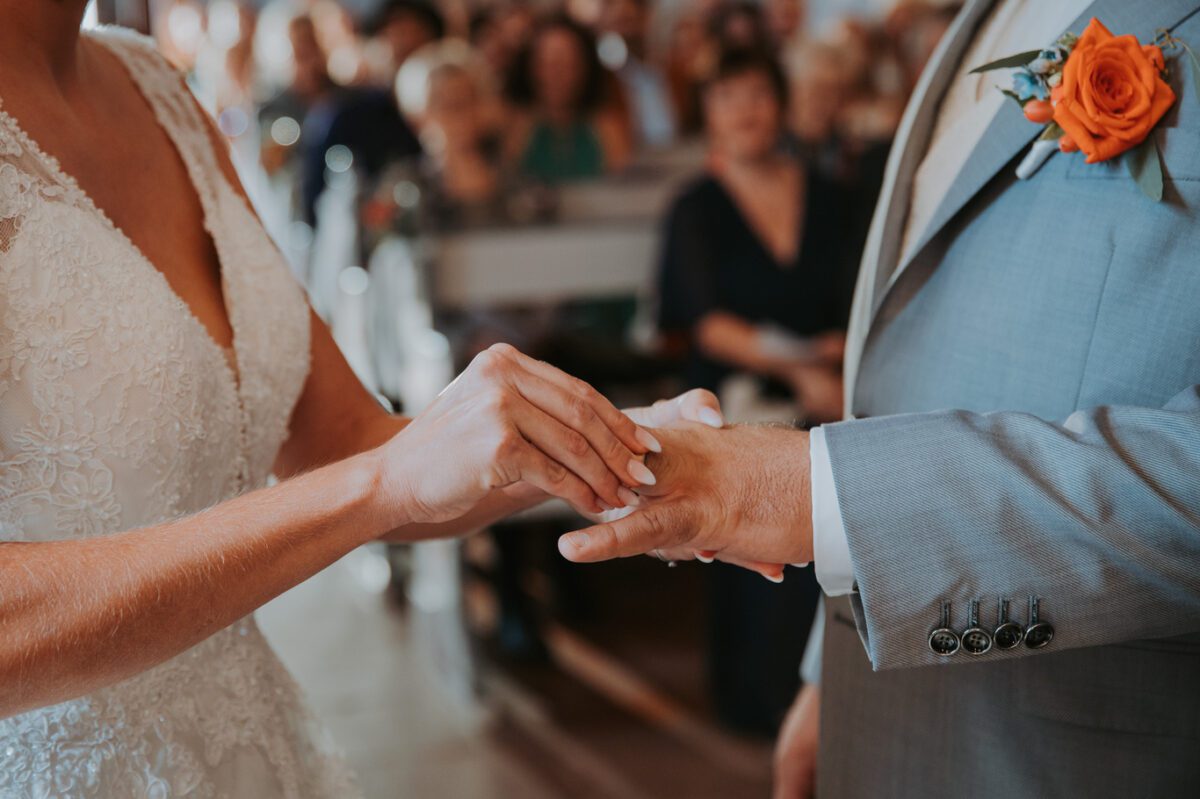 Bei einer standesamtlichen Trauung in Frankfurt steckt eine Braut in einem Spitzenkleid dem Bräutigam einen Ring an den Finger. Der Bräutigam, gekleidet in einen hellgrauen Anzug mit einer leuchtend orangefarbenen Rose als Ansteckblume, steht inmitten sanfter und warmer Beleuchtung. Im Hintergrund sitzen leicht verschwommene Gäste, die den intimen Moment des Fotoshootings verstärken. - Elis Wedding - Hochzeitsfotograf Frankfurt