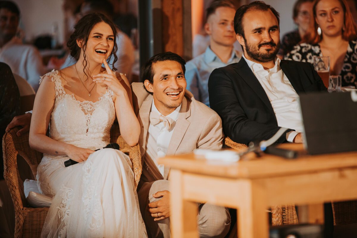 Eine Braut in einem Spitzenhochzeitskleid und ein Bräutigam in einem hellen Anzug sitzen lächelnd und lachend auf Korbstühlen. Neben ihnen sitzt ein Mann in einem dunklen Anzug. Das Trio, wunderschön eingefangen durch Fotografie, sieht sich etwas außerhalb des Kamerablickfelds auf einem Laptop an. Mehrere sitzende Gäste sind vor dem warm getönten Hintergrund verschwommen. - Elis Wedding - Hochzeitsfotograf Frankfurt