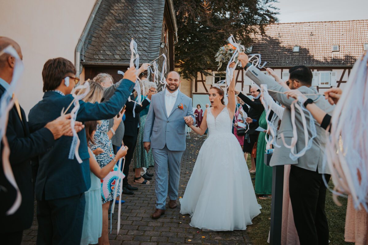 Eine Braut in einem weißen Brautkleid und ein Bräutigam in einem hellblauen Anzug gehen einen Gang entlang, der von Gästen gebildet wird, die weiße Bänder schwenken. Die Hand der Braut ist erhoben und hält einen Blumenstrauß. Die Szene spielt im Freien auf einem Kopfsteinpflasterweg, perfekt für ein Fotoshooting, flankiert von Gebäuden mit rustikalen Holzelementen. - Elis Wedding - Hochzeitsfotograf Frankfurt