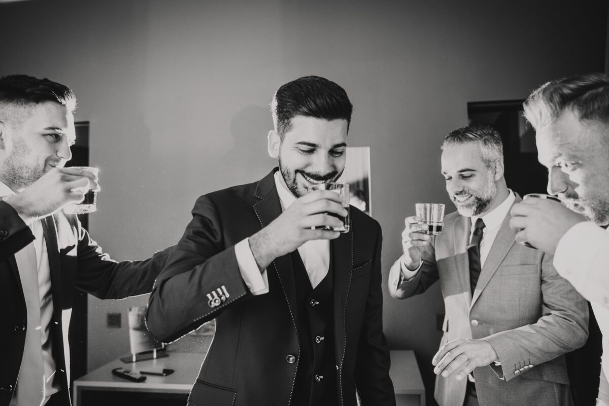 Ein Schwarzweißfoto zeigt vier Männer in formellen Anzügen, die ihre Gläser zum Anstoßen erheben. Der Mann in der Mitte lächelt und blickt auf sein Getränk. Sie wirken alle glücklich und mitten in der Feier, dicht beieinander in einem warm beleuchteten Raum – vielleicht aufgenommen von einem Top-Hochzeitsfotografen in Frankfurt während ihres Kennenlernens. - Elis Wedding - Hochzeitsfotograf Frankfurt