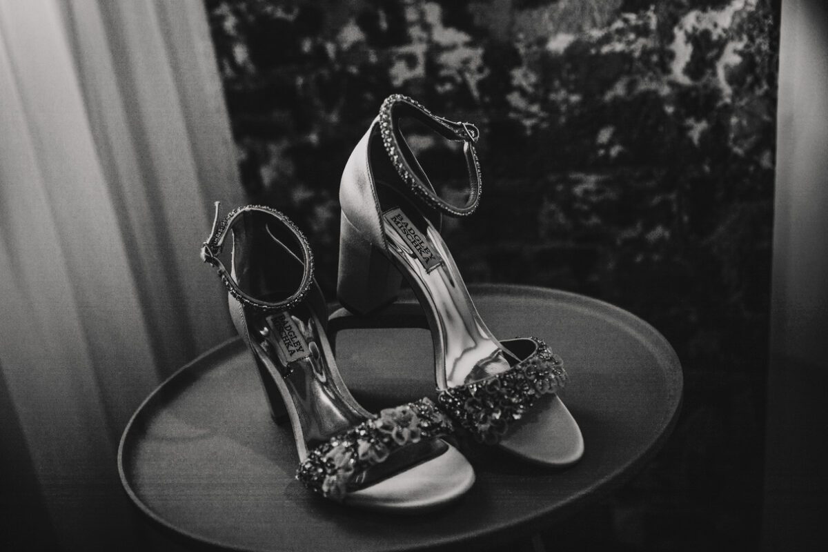 Ein Paar eleganter Stöckelschuhe steht auf einem kleinen runden Tisch. Die Schuhe haben mit Kristallen verzierte Knöchelriemen und die Zehenriemen sind mit aufwendigen Blumenverzierungen versehen. Dieses Schwarzweißfoto, das für einen Hochzeitsfotografen geeignet ist, hebt das glänzende, detaillierte Design der Schuhe vor einem dunklen, strukturierten Hintergrund hervor. - Elis Wedding - Hochzeitsfotograf Frankfurt