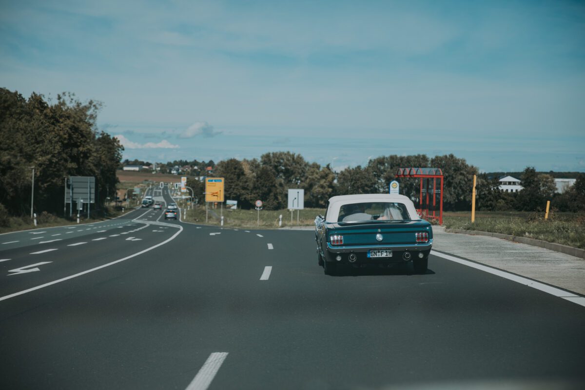 Ein blauer Oldtimer mit weißem Cabriodach fährt auf einer mehrspurigen Autobahn, umgeben von Grün, vielleicht auf dem Weg zu einem Fotoshooting oder einer standesamtlichen Trauung. Andere Autos teilen sich die Straße unter einem klaren blauen Himmel, während sich Ackerland und Bäume mit entfernten Gebäuden im Hintergrund erstrecken. - Elis Wedding - Hochzeitsfotograf Frankfurt
