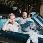 Braut und Bräutigam lächeln, als sie kurz nach ihrer standesamtlichen Trauung in einem blauen Oldtimer-Cabrio sitzen. Die Braut in weißem Kleid und Tiara winkt vom Beifahrersitz aus, während der Bräutigam in einem hellgrauen Anzug fährt. Diese wunderschöne Szene spielt sich vor üppigem Grün und gewölbten Fenstern in Frankfurt ab. - Elis Wedding - Hochzeitsfotograf Frankfurt