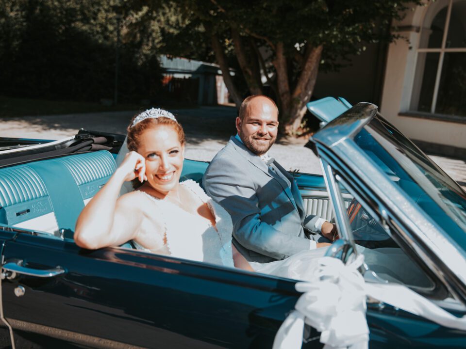 Braut und Bräutigam lächeln, als sie kurz nach ihrer standesamtlichen Trauung in einem blauen Oldtimer-Cabrio sitzen. Die Braut in weißem Kleid und Tiara winkt vom Beifahrersitz aus, während der Bräutigam in einem hellgrauen Anzug fährt. Diese wunderschöne Szene spielt sich vor üppigem Grün und gewölbten Fenstern in Frankfurt ab. - Elis Wedding - Hochzeitsfotograf Frankfurt