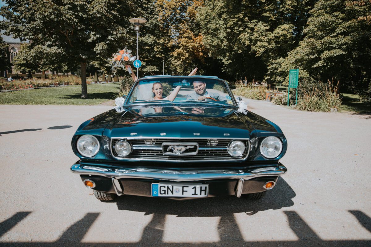Ein blauer Ford Mustang Cabriolet steht auf einem sonnenbeschienenen Weg. Zwei lächelnde Personen sitzen auf den Vordersitzen und heben freudig ihre Arme, als würden sie für ein reizendes Fotoshooting posieren. Im Hintergrund sind grüne Bäume und ein gepflegter Garten zu sehen, was auf eine entspannte und fröhliche Atmosphäre hindeutet. Auf dem Nummernschild des Autos steht „GN-F1H“. - Elis Wedding - Hochzeitsfotograf Frankfurt