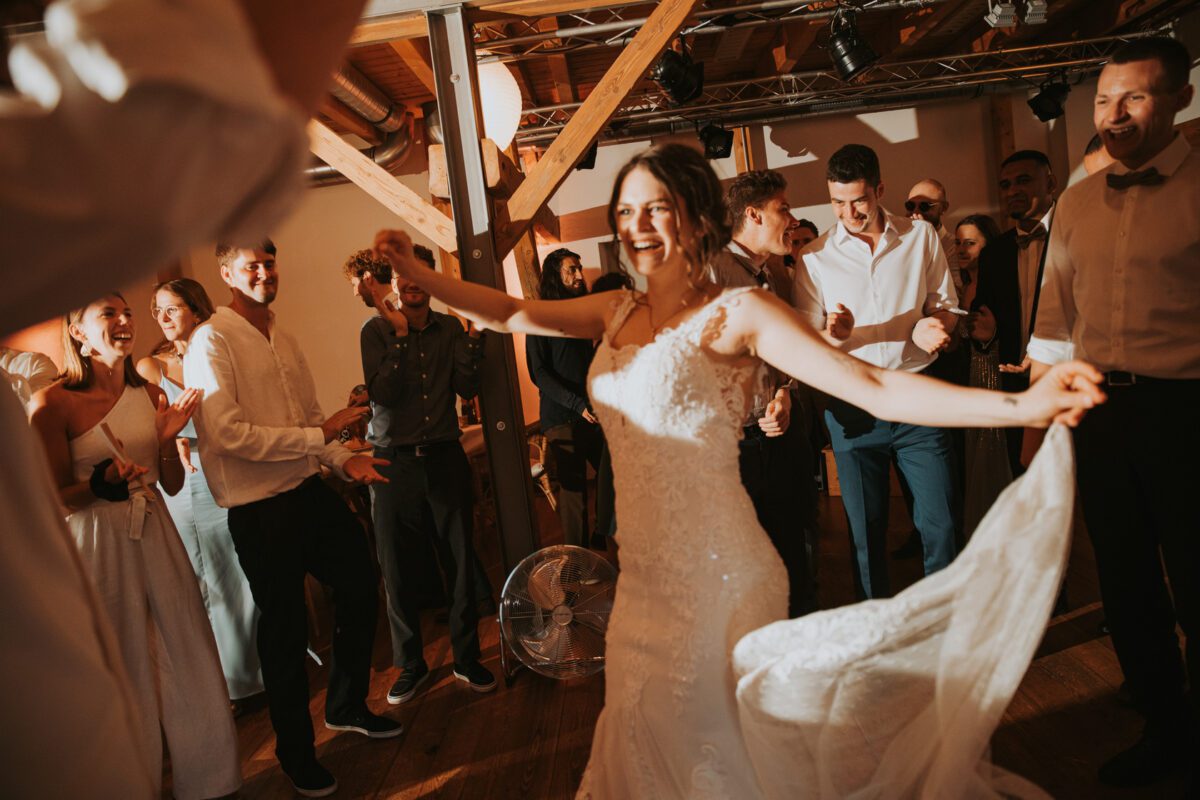Eine Braut in einem weißen Spitzenbrautkleid tanzt fröhlich auf ihrer Hochzeitsfeier in Frankfurt. Sie ist von einer lebhaften Gruppe von Gästen umgeben, die in eleganter Freizeitkleidung gekleidet sind und klatschen und jubeln. Der rustikale Innenbereich mit Holzbalken und sanfter Beleuchtung trägt zur festlichen Atmosphäre bei, perfekt für ein Fotoshooting. - Elis Wedding - Hochzeitsfotograf Frankfurt
