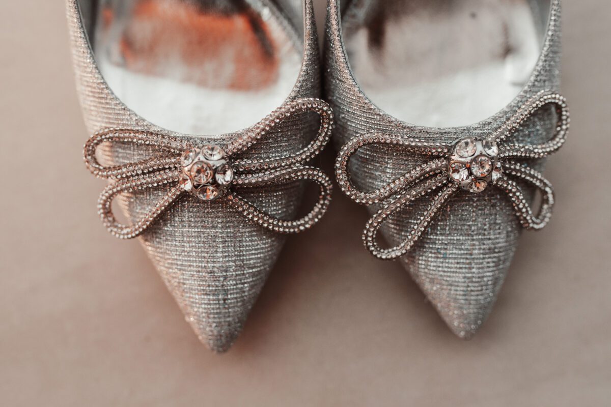 Nahaufnahme eines Paars eleganter, silberfarbener High Heels mit spitzer Spitze und aufwendig verzierten Perlenschleifen und Strasssteinen auf der Vorderseite. Der Stoff der Schuhe hat eine schimmernde, strukturierte Optik, die ihnen einen glamourösen und anspruchsvollen Look verleiht – perfekt als Hintergrund für ein Hochzeitsfoto auf einer hellen, neutralen Oberfläche. - Elis Wedding - Hochzeitsfotograf Frankfurt
