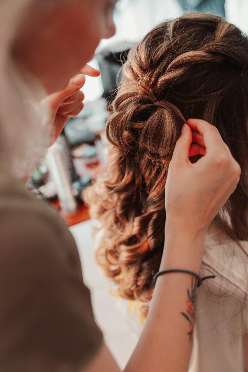 Eine Person mit lockigem braunem Haar sitzt, während eine andere Person es zu einer komplizierten Hochsteckfrisur mit Zopf und Locken frisiert. Der Stylist, der sich möglicherweise auf eine standesamtliche Trauung vorbereitet, trägt ein Armband und hat ein Tattoo auf dem Unterarm. Die Szene spielt im Innenbereich, im Hintergrund sind verschiedene Haarstyling-Tools zu sehen, die einen perfekten Moment für Hochzeitsfotografen festhalten. - Elis Wedding - Hochzeitsfotograf Frankfurt