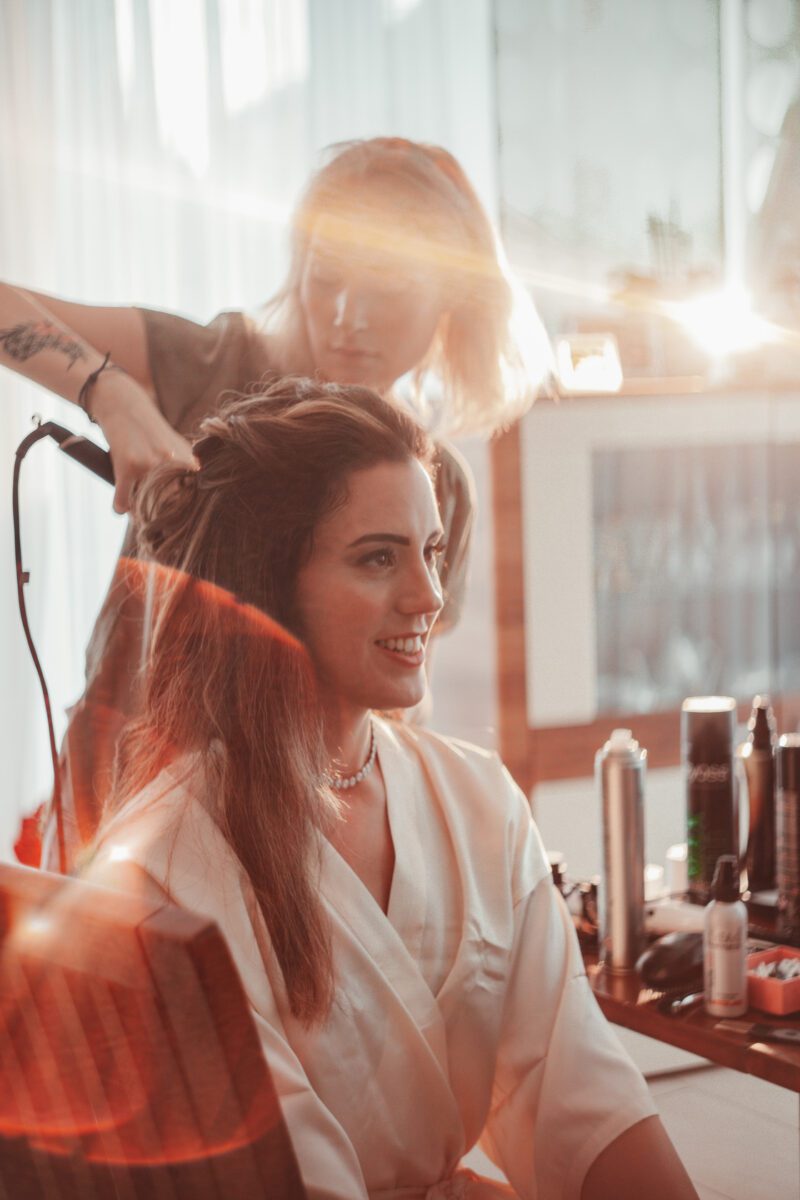 Eine Frau in einem weißen Morgenmantel sitzt lächelnd auf einem Stuhl, während ein Friseur hinter ihr ihr Haar mit einem Lockenstab frisiert. Die Szene ist warm beleuchtet, natürliches Licht fällt ein und hebt mehrere Haarprodukte auf dem Tisch in der Nähe hervor. Aufgenommen von einem Hochzeitsfotografen in Frankfurt, verleihen Linsenreflexionen dem Bild einen verträumten Effekt. - Elis Wedding - Hochzeitsfotograf Frankfurt