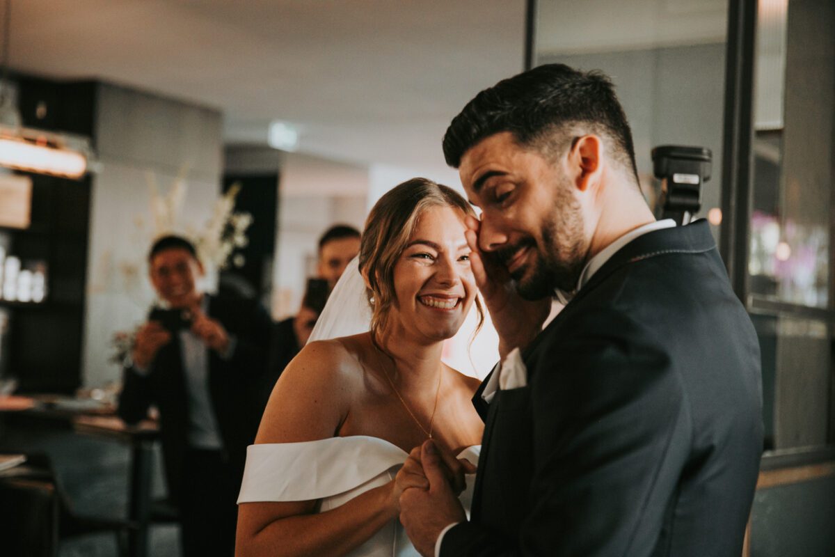 Braut und Bräutigam in Hochzeitskleidung teilen einen freudigen Moment im Haus während ihres Kennenlernens. Die Braut, die ein weißes schulterfreies Kleid trägt, lächelt strahlend, während sie die Hand des Bräutigams hält. Der Bräutigam, in einem dunklen Anzug, hat einen verspielten Gesichtsausdruck. Im Hintergrund lächeln zwei Menschen und halten den Moment mit Fotos fest. - Elis Wedding - Hochzeitsfotograf Frankfurt