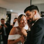 Braut und Bräutigam in Hochzeitskleidung teilen einen freudigen Moment im Haus während ihres Kennenlernens. Die Braut, die ein weißes schulterfreies Kleid trägt, lächelt strahlend, während sie die Hand des Bräutigams hält. Der Bräutigam, in einem dunklen Anzug, hat einen verspielten Gesichtsausdruck. Im Hintergrund lächeln zwei Menschen und halten den Moment mit Fotos fest. - Elis Wedding - Hochzeitsfotograf Frankfurt