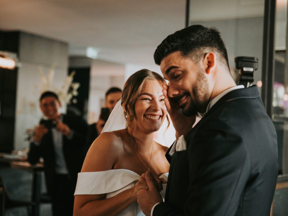 Braut und Bräutigam in Hochzeitskleidung teilen einen freudigen Moment im Haus während ihres Kennenlernens. Die Braut, die ein weißes schulterfreies Kleid trägt, lächelt strahlend, während sie die Hand des Bräutigams hält. Der Bräutigam, in einem dunklen Anzug, hat einen verspielten Gesichtsausdruck. Im Hintergrund lächeln zwei Menschen und halten den Moment mit Fotos fest. - Elis Wedding - Hochzeitsfotograf Frankfurt