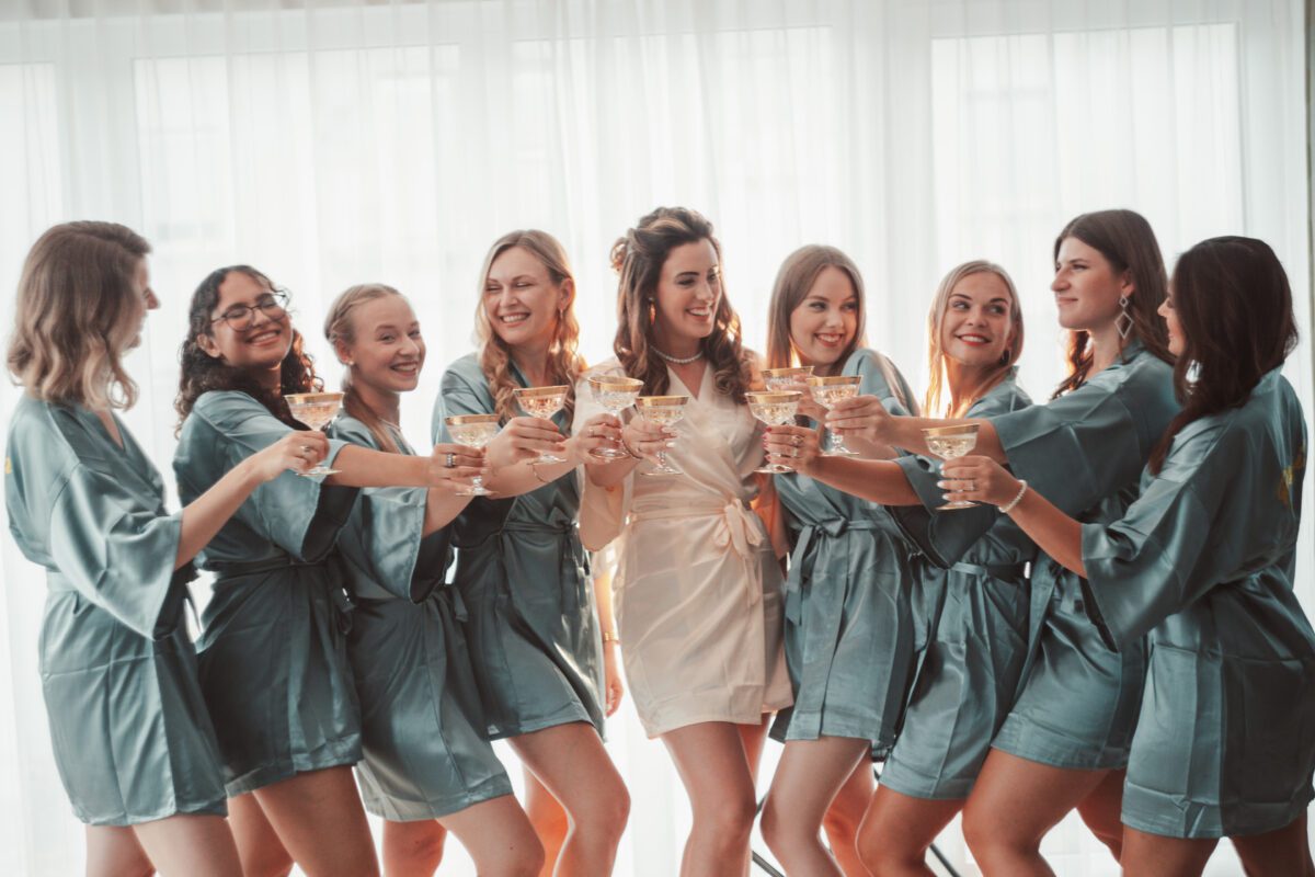 Eine Braut in einem weißen Gewand steht in der Mitte einer Gruppe von sieben Frauen in passenden grünen Gewändern. Sie sind drinnen, lächeln und erheben Champagnergläser zu einem festlichen Toast. Der erfahrene Hochzeitsfotograf fängt diesen strahlenden Moment ein, wobei hauchdünne weiße Vorhänge einen weichen Hintergrund bilden. - Elis Wedding - Hochzeitsfotograf Frankfurt