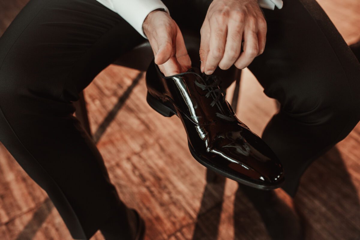 Eine Person in weißem Hemd und schwarzer Hose sitzt da und bindet die Schnürsenkel eines glänzenden schwarzen Abendschuhs. Der Fokus liegt auf den Händen und dem Schuh, der das Licht reflektiert. Der Hintergrund besteht aus einem Holzboden mit einem warmen, weichen Lichteffekt, der einen Moment vor ihrer standesamtlichen Trauung in Frankfurt einfängt. - Elis Wedding - Hochzeitsfotograf Frankfurt